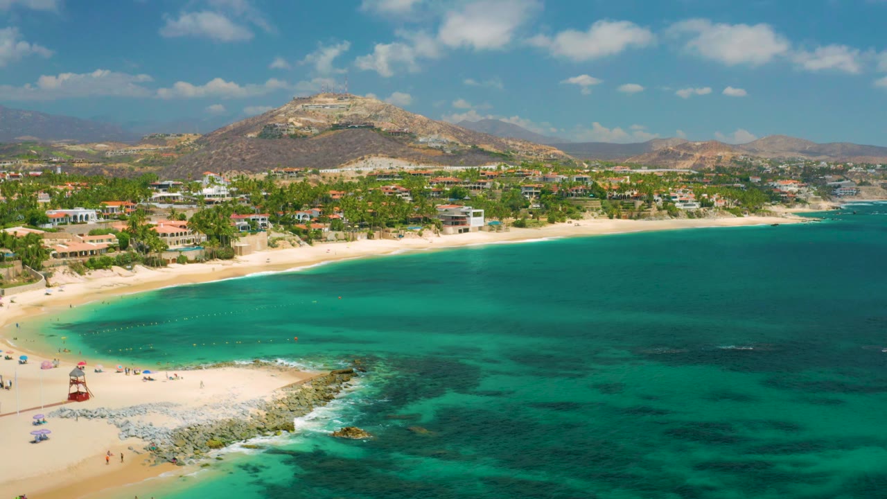 Palmilla Beach Cabo is seen with the rocky point lower left, a line of beach umbrellas, visible tiny beachgoers, bright turquoise shallows, and low-rise buildings beneath dry hills.