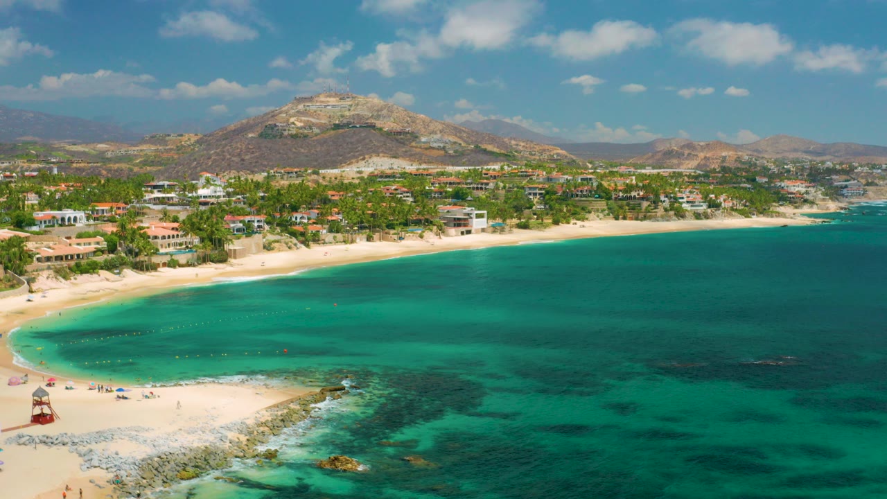 Opening aerial frame over Palmilla Beach Cabo, Mexico, with turquoise water in the foreground, a pale sand curve at left, beach buildings and palms behind it, hills on the horizon, and no visible people.