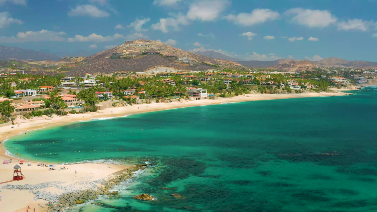 At 0:01, the Palmilla Beach Cabo aerial view holds on the broad green-blue bay, with the sandy beach running across the left and center, coastal buildings behind the palms, distant hills, and no visible people.