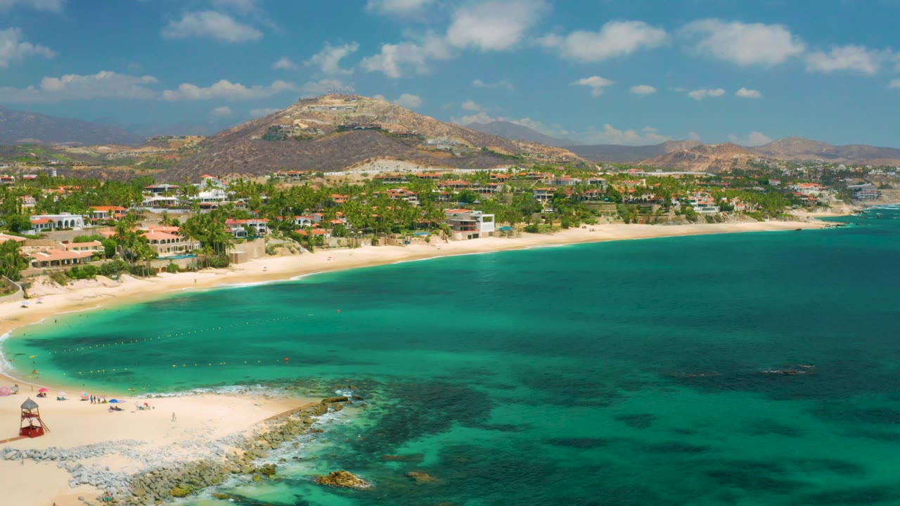 At 0:02, clear turquoise water fills more of the foreground while Palmilla Beach Cabo, Mexico remains visible along the left shore with beach structures, palms, low buildings, hills, and no visible people.
