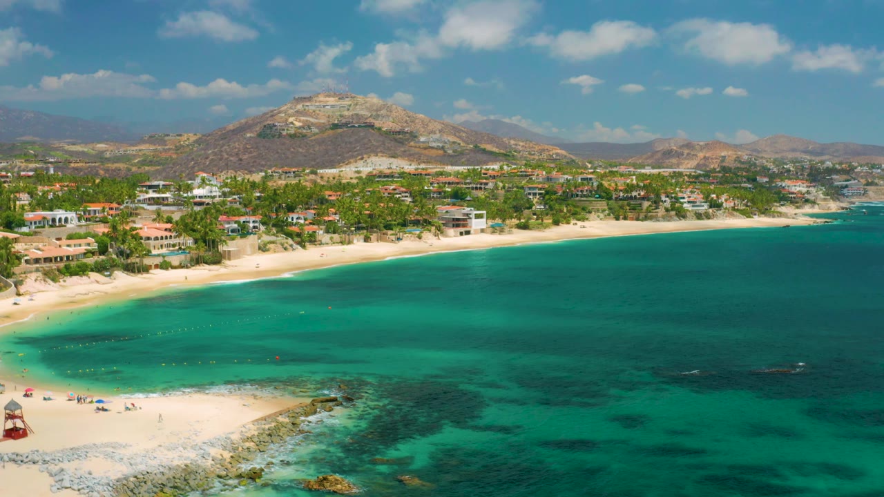 Aerial shoreline with turquoise sea, sand, rocky edge, homes, palms, and hills.