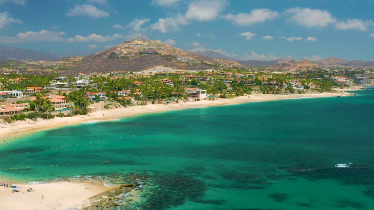 Aerial frame at Palmilla Beach Cabo, Mexico showing turquoise water in the foreground, pale sand and beachfront buildings beyond it, arid hills under a blue sky, and no visible people.