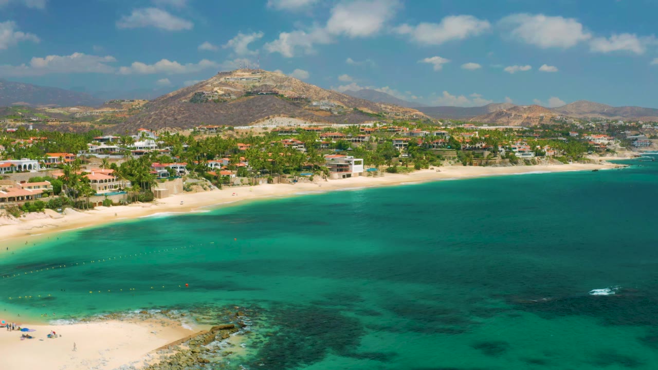 Wide aerial view at Palmilla Beach Cabo, Mexico with shallow turquoise water, a pale beach, palm-lined shoreline buildings, rocky hills in back, and no visible people.