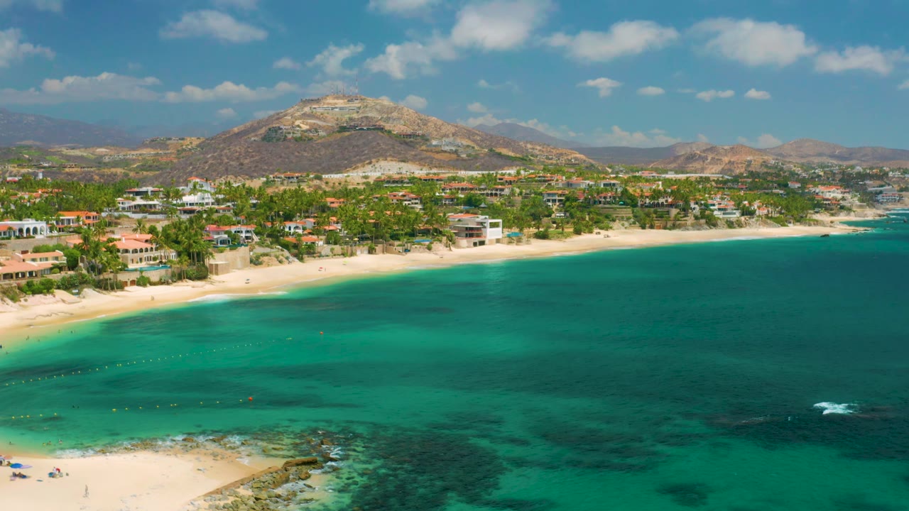 Aerial view at Palmilla Beach Cabo, Mexico showing a broad turquoise bay, sandy shoreline with palms and low buildings, dry hills behind, and no visible people.