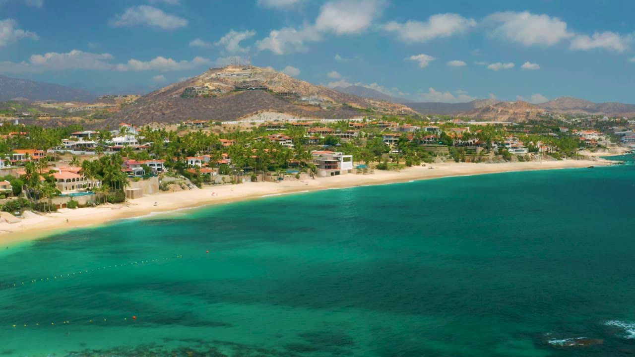 Wide aerial coastline with green-blue water, beach houses, palms, and hills.