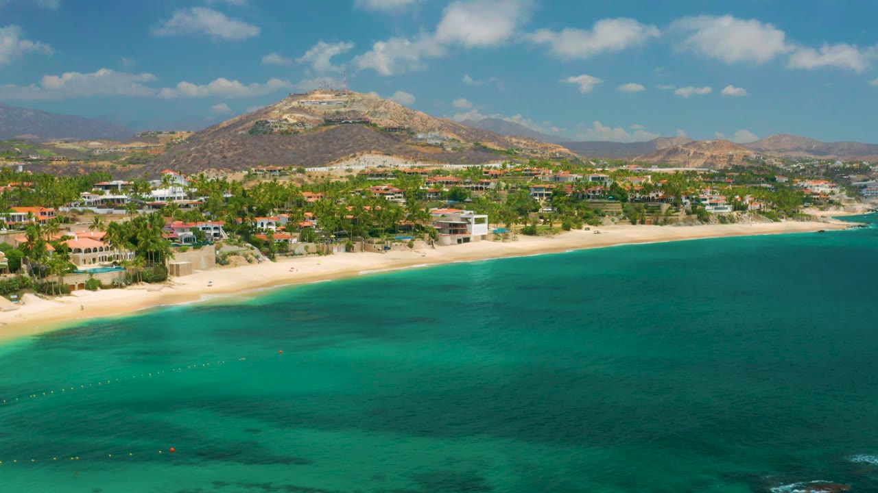 Palmilla Beach Cabo, Mexico appears from above with turquoise bay water, a narrow sand strip, residential buildings, a rocky mountain, and no visible people.