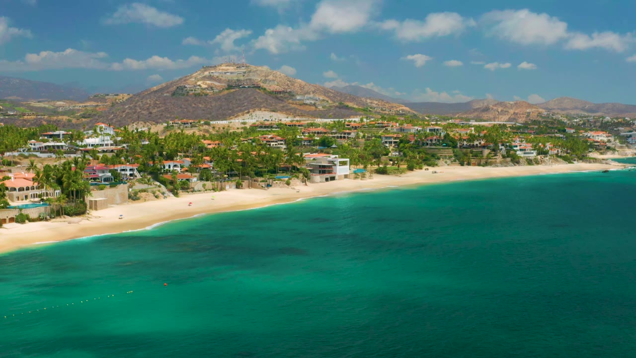 Aerial frame over Palmilla Beach Cabo, Mexico, showing clear green water across the lower frame, a curved sandy beach, palms and low homes, distant hills, and no visible people.
