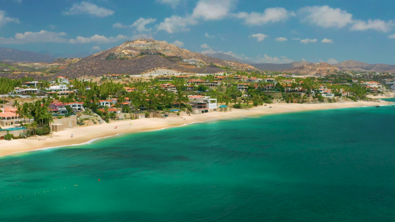 Wide aerial frame of Palmilla Beach Cabo, Mexico, with teal sea in the foreground, a bright sand strip, clustered coastal homes, palms, dry hillside terrain, and no visible people.
