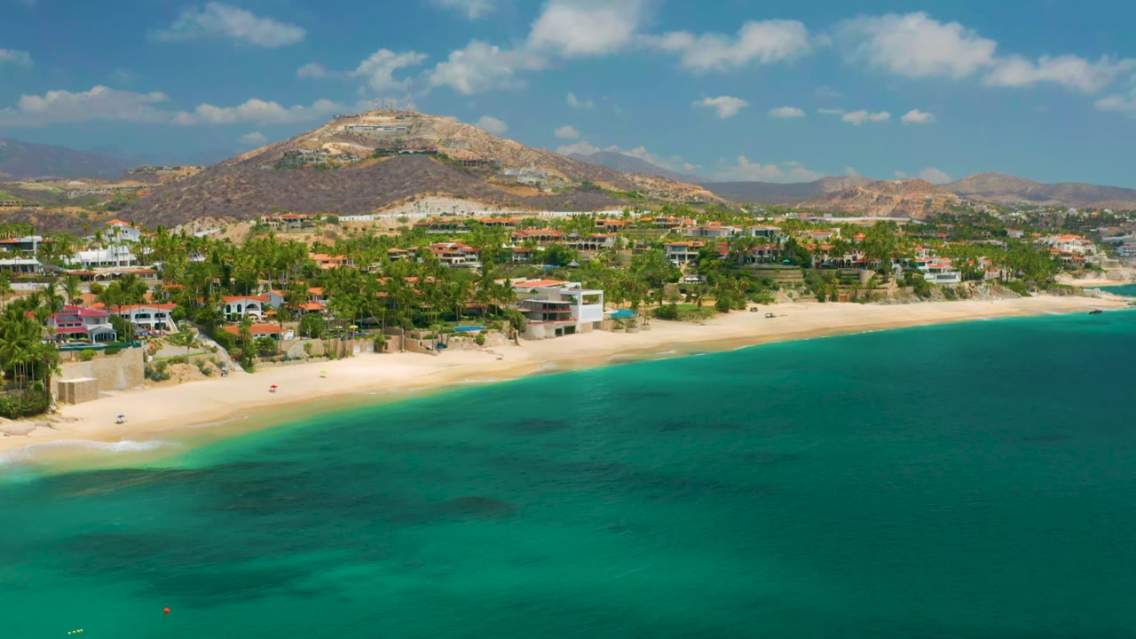 Palmilla Beach Cabo coastline from above, showing teal water in front, a pale sand strip, shorefront buildings among palms, layered hills in the distance, and no visible people.