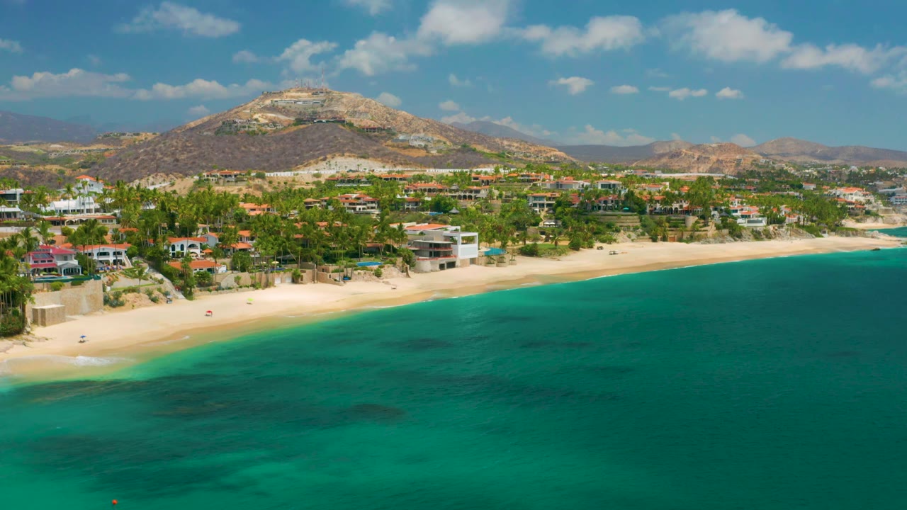 Aerial view at Palmilla Beach Cabo, Mexico with blue-green ocean filling the lower frame, beach and low buildings along the shore, arid hills beyond town, and no visible people.