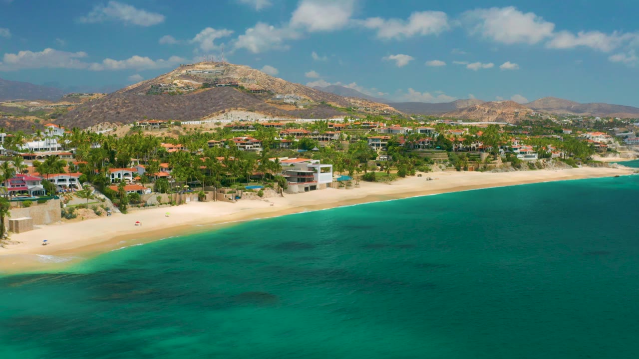 Aerial beach scene with shoreline homes, palms, turquoise sea, and mountains