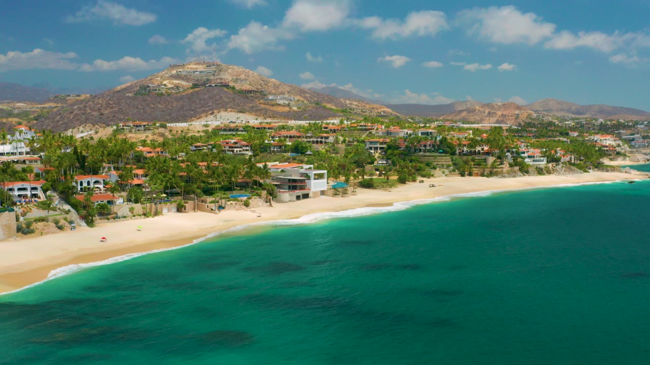 Opening aerial frame over Palmilla Beach Cabo, Mexico, with turquoise water in front, pale sand, beachfront homes, palms, dry hills, scattered clouds, and no visible people.