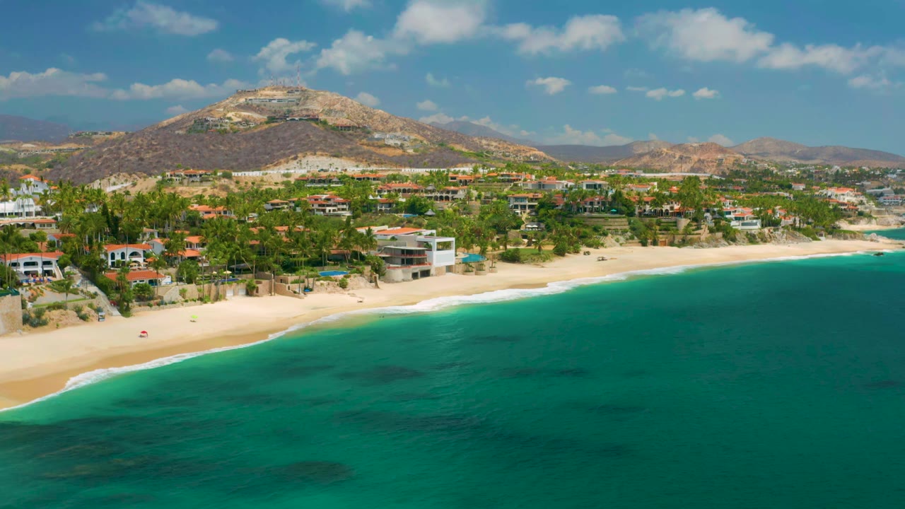Palmilla Beach Cabo aerial view a second into the clip, showing more open turquoise water in the foreground, a curved sand edge, homes behind palms, dry hills, and no visible people.