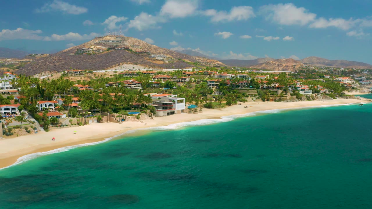 Wide aerial frame over Palmilla Beach Cabo with beach houses and palms spanning the midground, clear turquoise water below, dry hills behind, small clouds above, and no visible people.