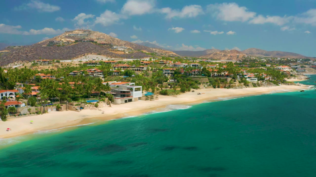 Aerial coastline view with clear green-blue water, beach, palms, homes, and hills.