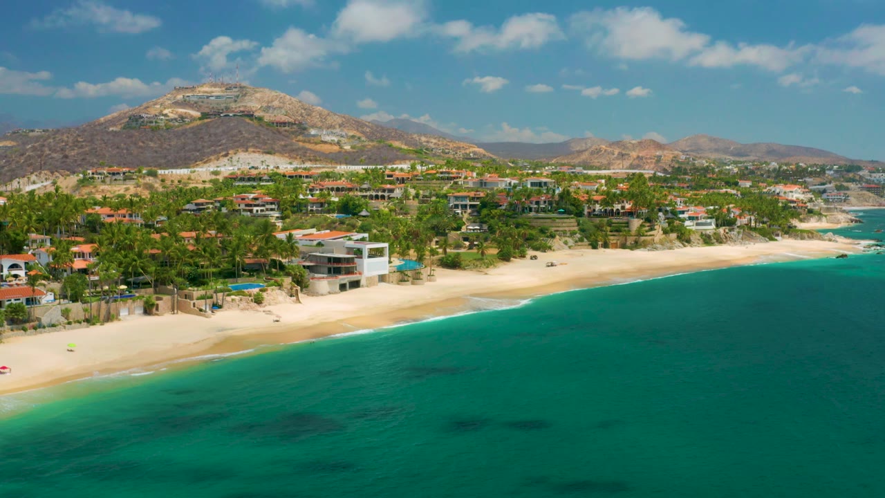 Palmilla Beach Cabo aerial frame with the ocean occupying the lower half, a band of sand and homes through the center, mountains across the background, and no visible people.