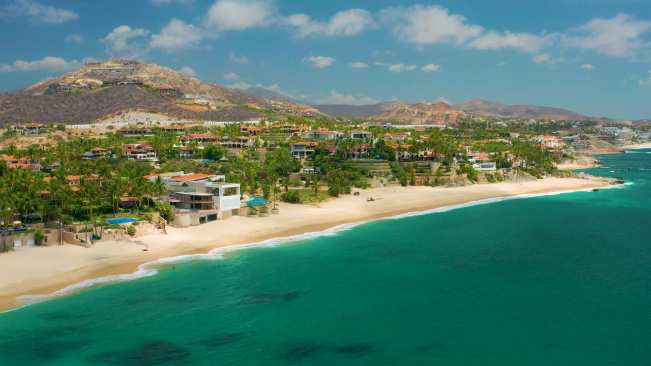 Opening frame over Palmilla Beach Cabo, Mexico, with turquoise sea in the foreground, pale sand and beachfront homes along the shore, dry mountains beyond, and no visible people.