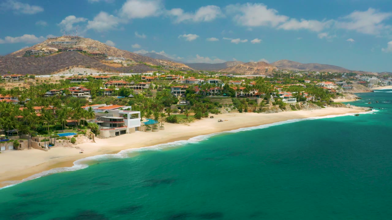 Aerial view over Palmilla Beach Cabo with more open water filling the lower frame, beachfront homes and palms along the sand, rugged hills behind them, and no visible people.
