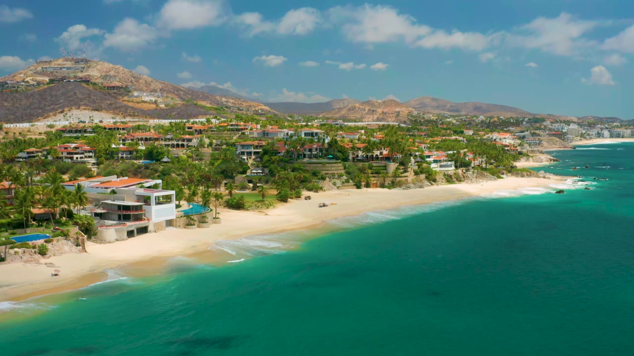 Palmilla Beach Cabo frame showing a modern white beach house near the lower left, palm trees and homes along the shore, turquoise sea to the right, hills beyond, and no visible people.