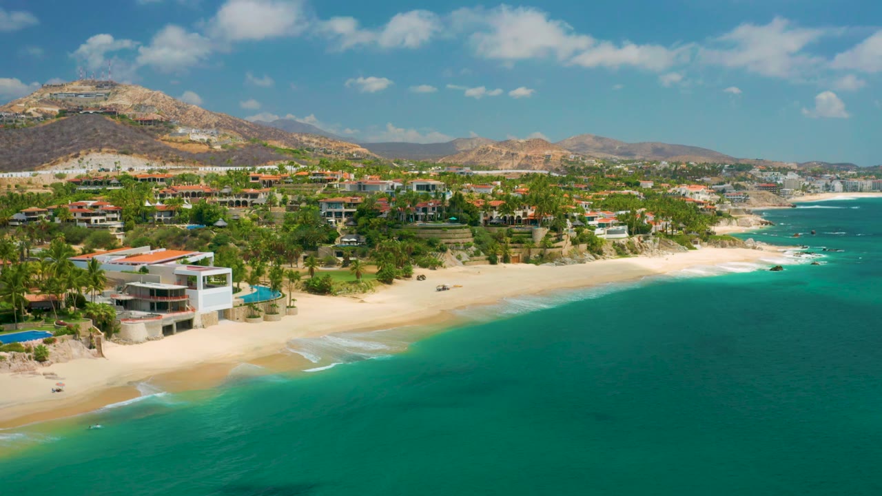 Palmilla Beach Cabo wide aerial with more open water on the right, a curved sandy beach through the center, hillside homes in the midground, distant mountains, and no visible people.