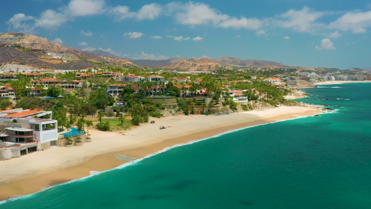 Opening aerial frame over Palmilla Beach Cabo, Mexico, with turquoise water, sandy shoreline, hillside buildings, dry mountains, scattered clouds, and no visible people.