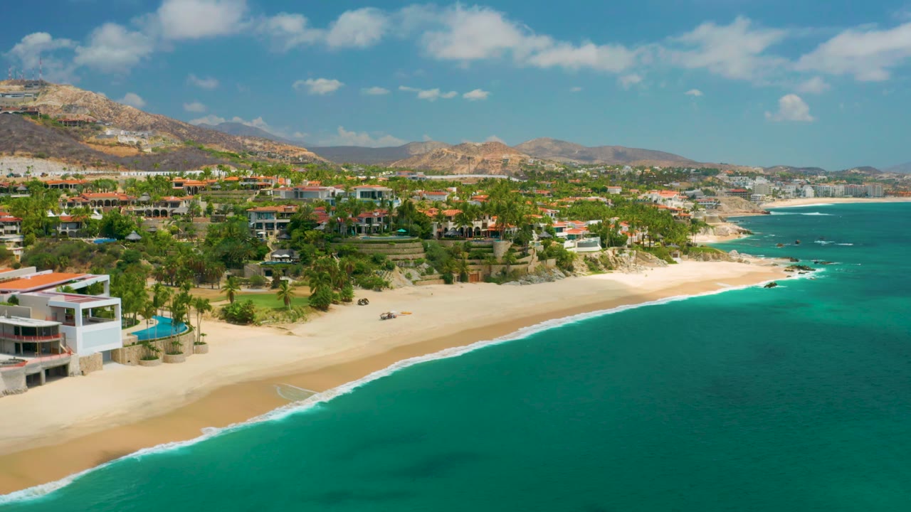 Aerial view of Palmilla Beach Cabo, Mexico, with palm trees and hillside homes above a pale sandy beach, calm turquoise ocean filling the right side, and no visible people.
