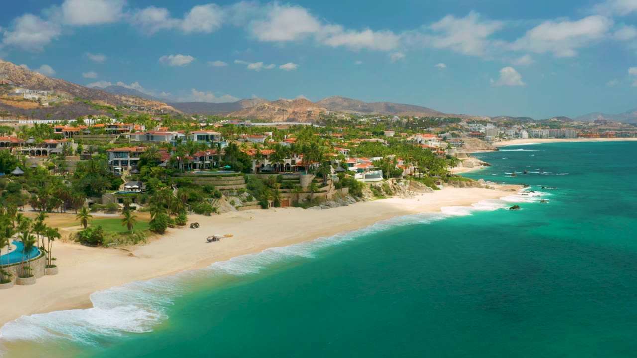 Wide aerial beach view with waves, palm trees, villas, town, and mountains.