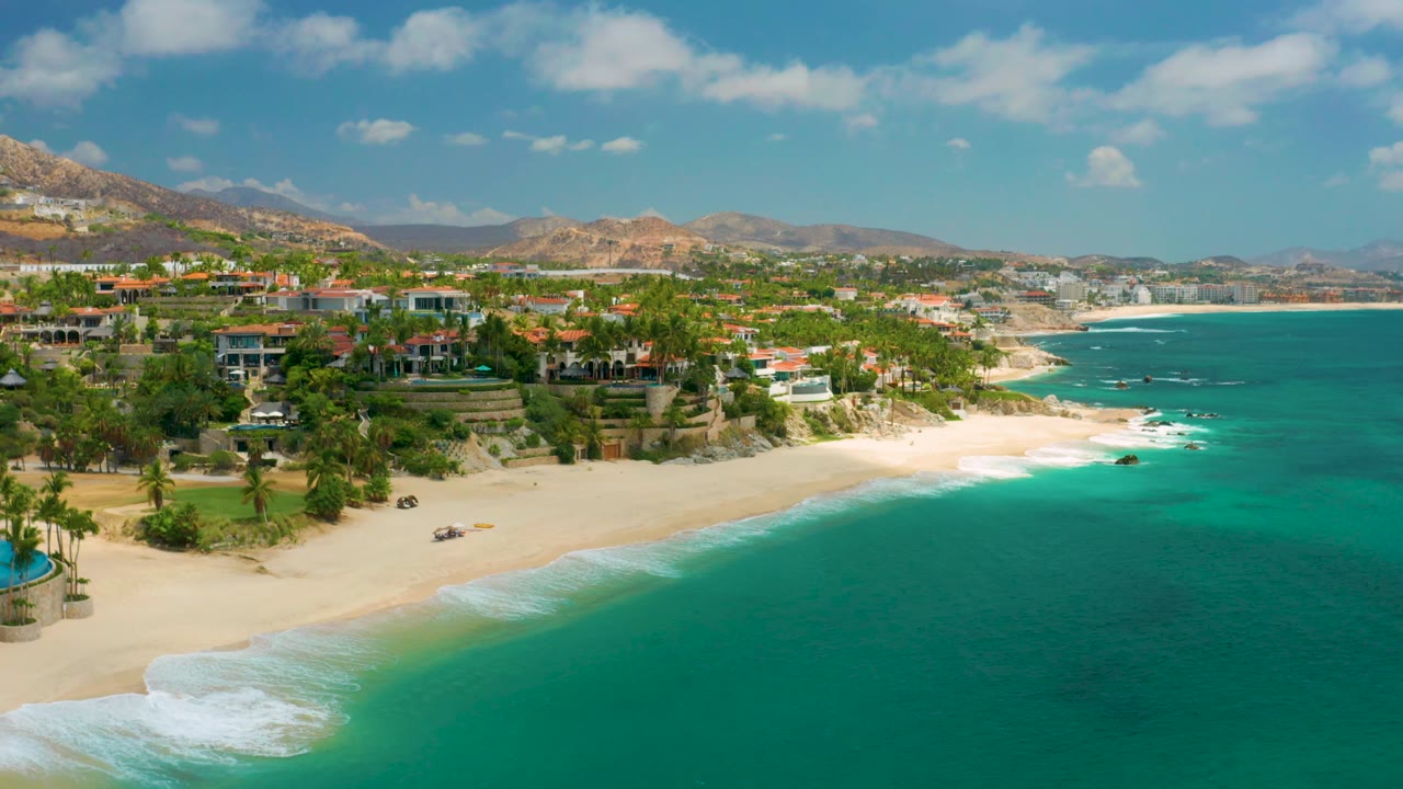 Palmilla Beach Cabo, Mexico coastline from above, with the beach curve more centered, green-blue water at right, hillside homes and palms at left, and no visible people.