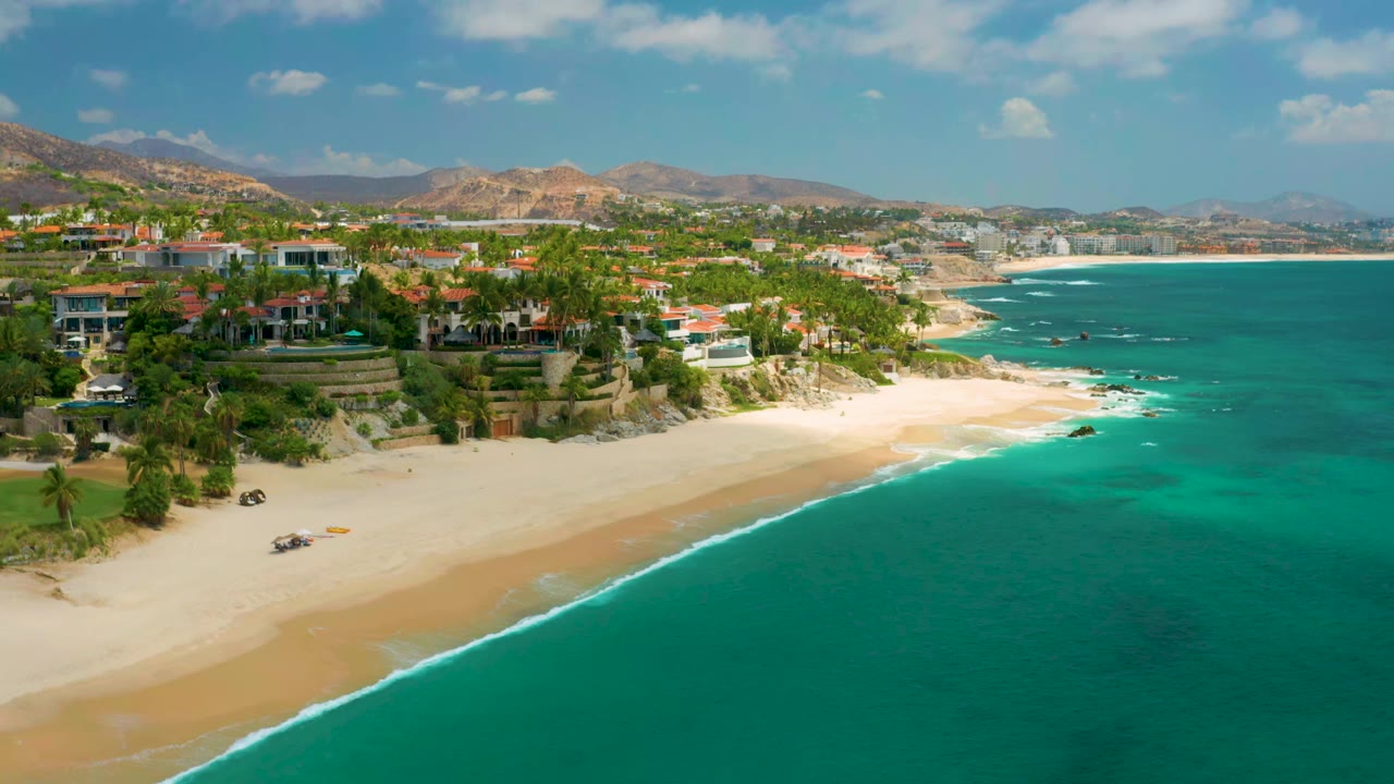 Wide aerial coastline with surf line, beach homes, and turquoise water.