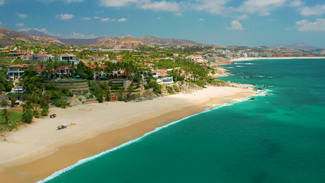 Palmilla Beach Cabo aerial frame with more open water at right, a curving sandy beach at left, hillside residences, palms, low mountains, and no visible people.