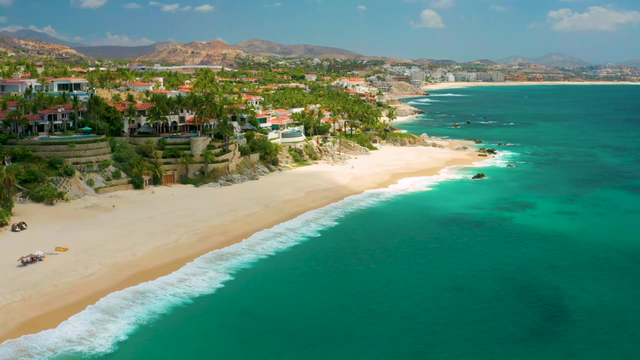 Aerial coastline with surf, beach homes or resort buildings, palms, and blue-green sea.