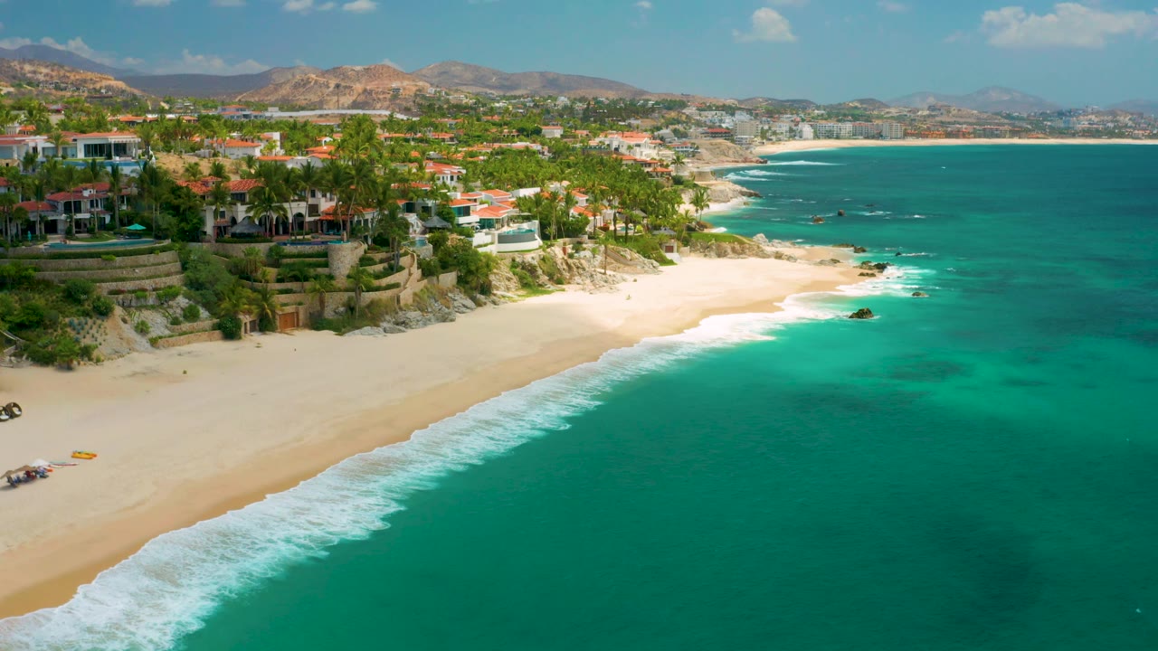 Palmilla Beach Cabo, Mexico appears in a high shoreline view with more open water to the right, the surf line running diagonally across the sand, coastal buildings to the left, and no visible people.