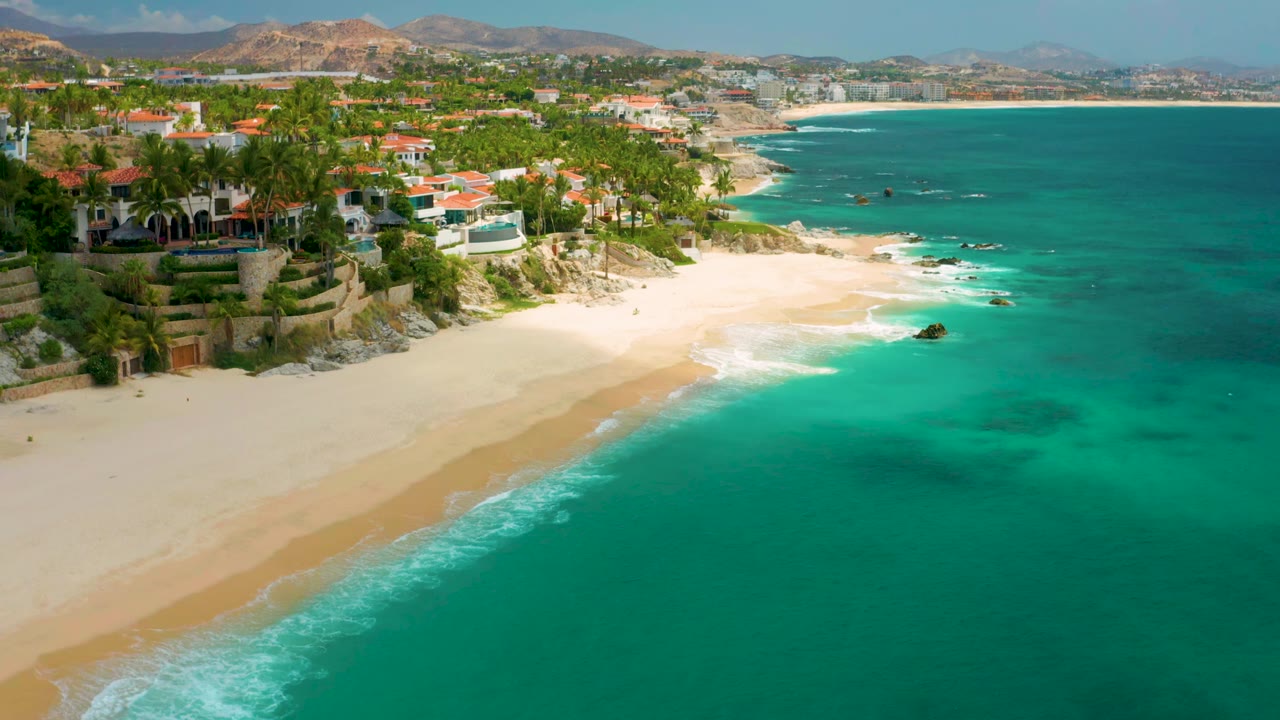 Aerial view along Palmilla Beach Cabo with the shoreline running diagonally, hotel buildings and palms on the left, blue-green water on the right, and no visible people.