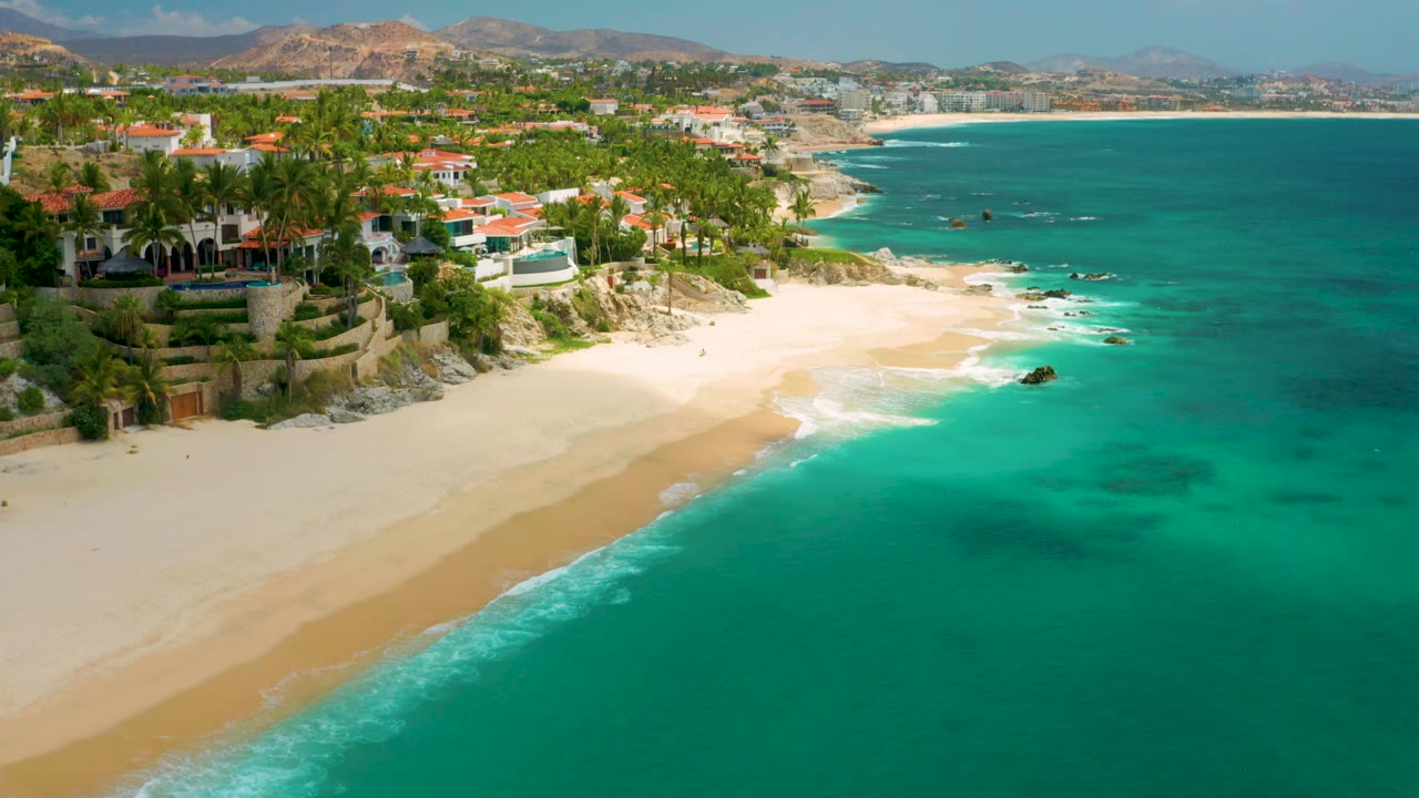 Palmilla Beach Cabo aerial frame showing a broad sweep of sand and water, with resort landscaping and red-roofed buildings at left and low mountains in the background, no visible people.