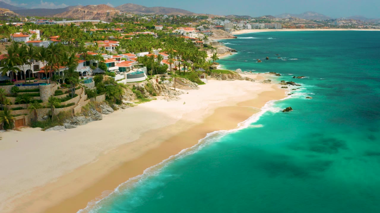 Opening aerial frame over Palmilla Beach Cabo, Mexico, with turquoise water, pale sand, palm trees, red-roofed homes, distant hills, and no visible people.