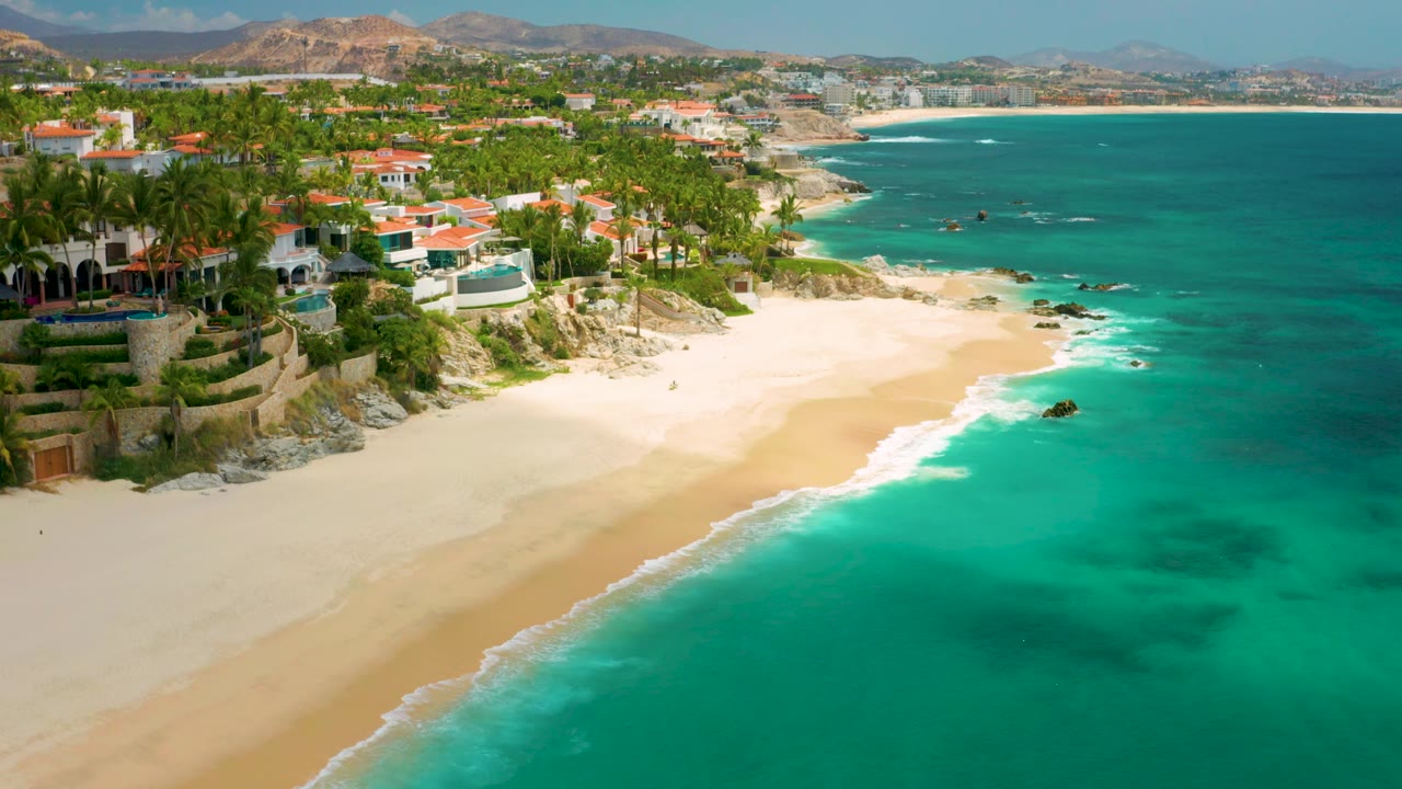 Palmilla Beach Cabo aerial frame with bright turquoise water filling the right side, palm-lined homes on the left, and no visible people on the sand.