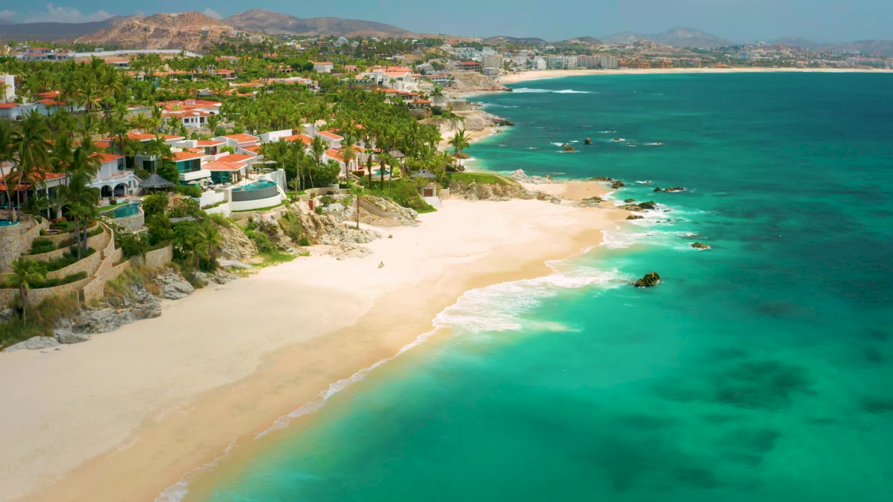 Aerial view at 0:01 over Palmilla Beach Cabo, Mexico, showing the beach curving below palm-covered homes, clear shallow water, darker reef patches, distant hills, and no visible people.