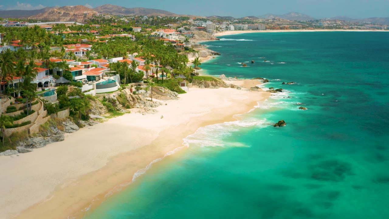 Aerial frame from Palmilla Beach Cabo, Mexico, with red-roof and white beachfront homes on the left, pale sand below, blue-green ocean across the right side, and no visible people.