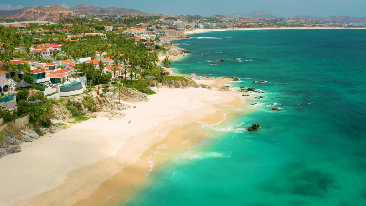 Aerial view over Palmilla Beach Cabo, Mexico, with pale sand at lower left, aquamarine water at right, red-roofed beachfront homes and palms along the bluff, and a small child visible near the shoreline.