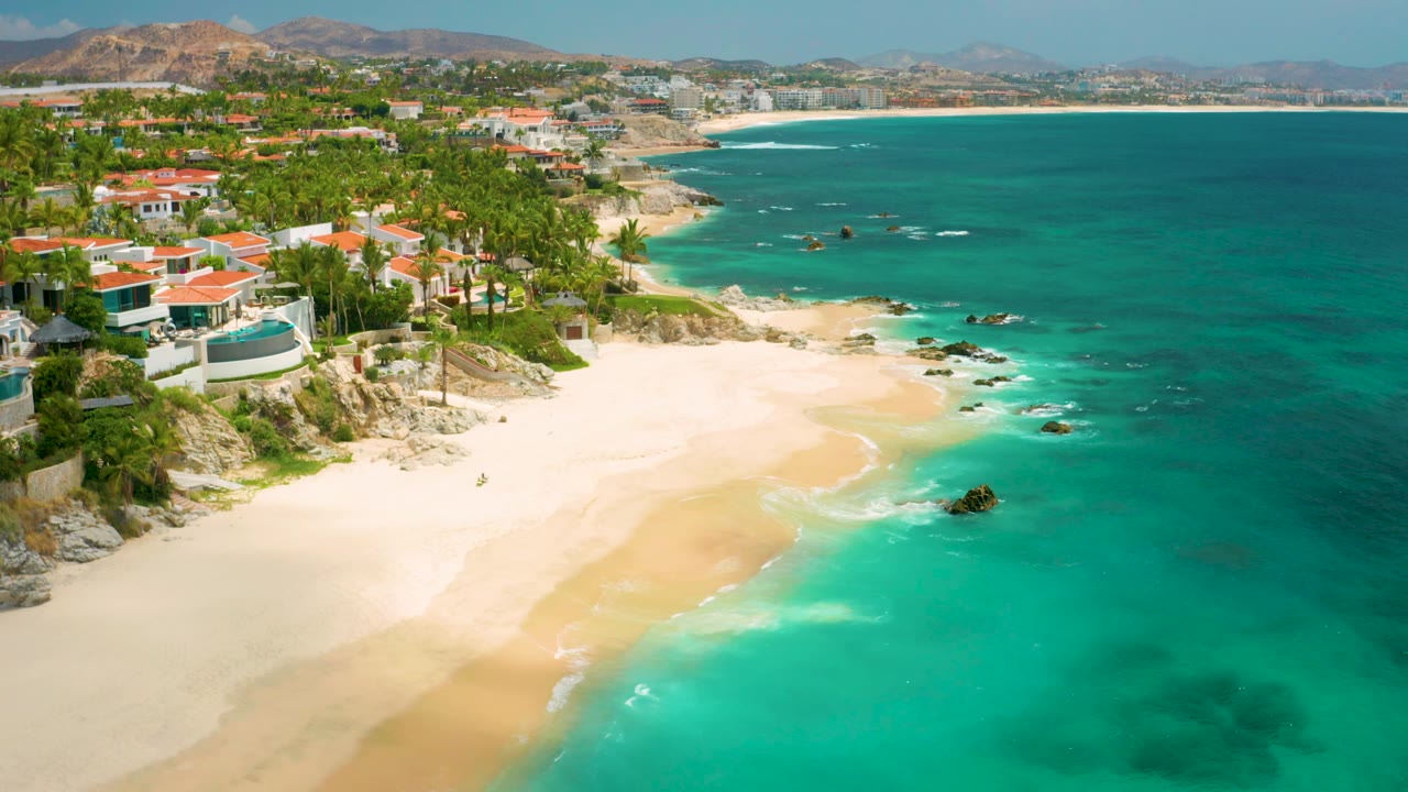 High view of Palmilla Beach Cabo with the beach widening in the foreground, white foam tracing the waterline, rocks offshore, homes and palms at left, and a small child still visible as a tiny figure on the sand.
