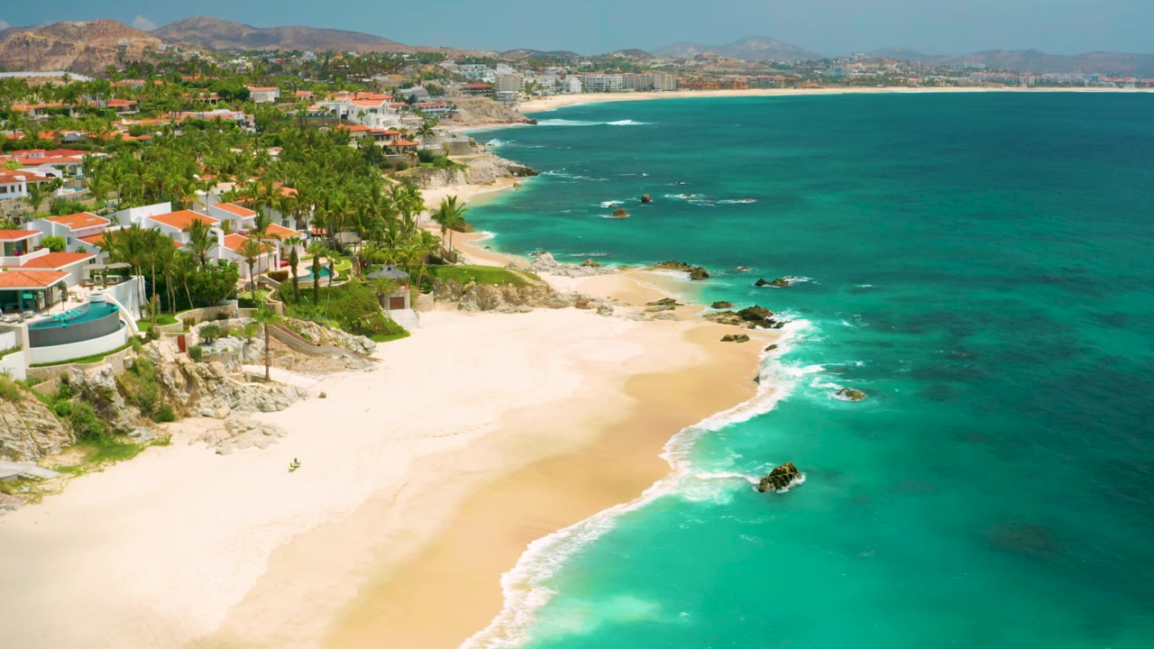 Wide aerial shoreline view with homes, palms, sand, surf, and blue-green water.
