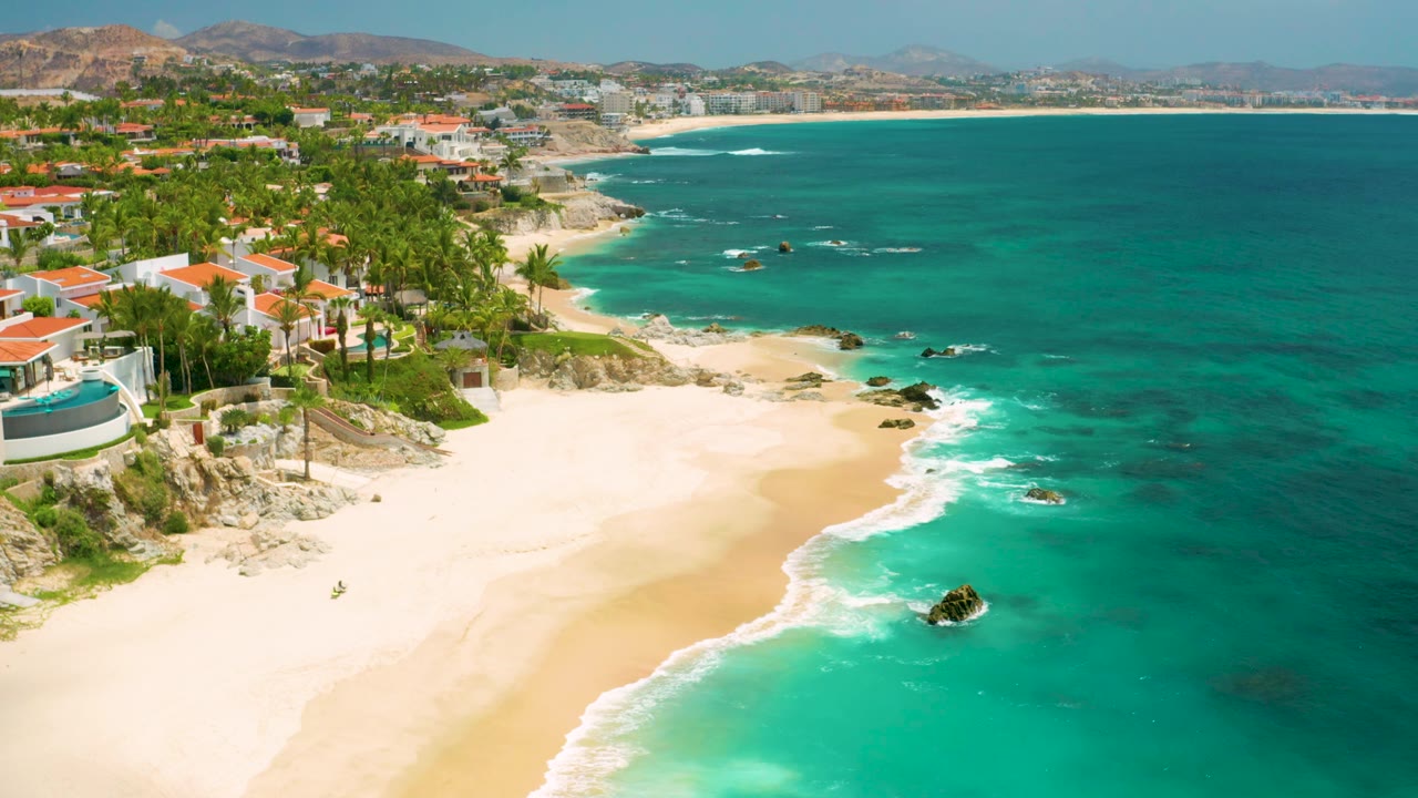 Palmilla Beach Cabo appears from above with red-roof homes on the left, a broad empty sand area, darker offshore patches, and no visible people.