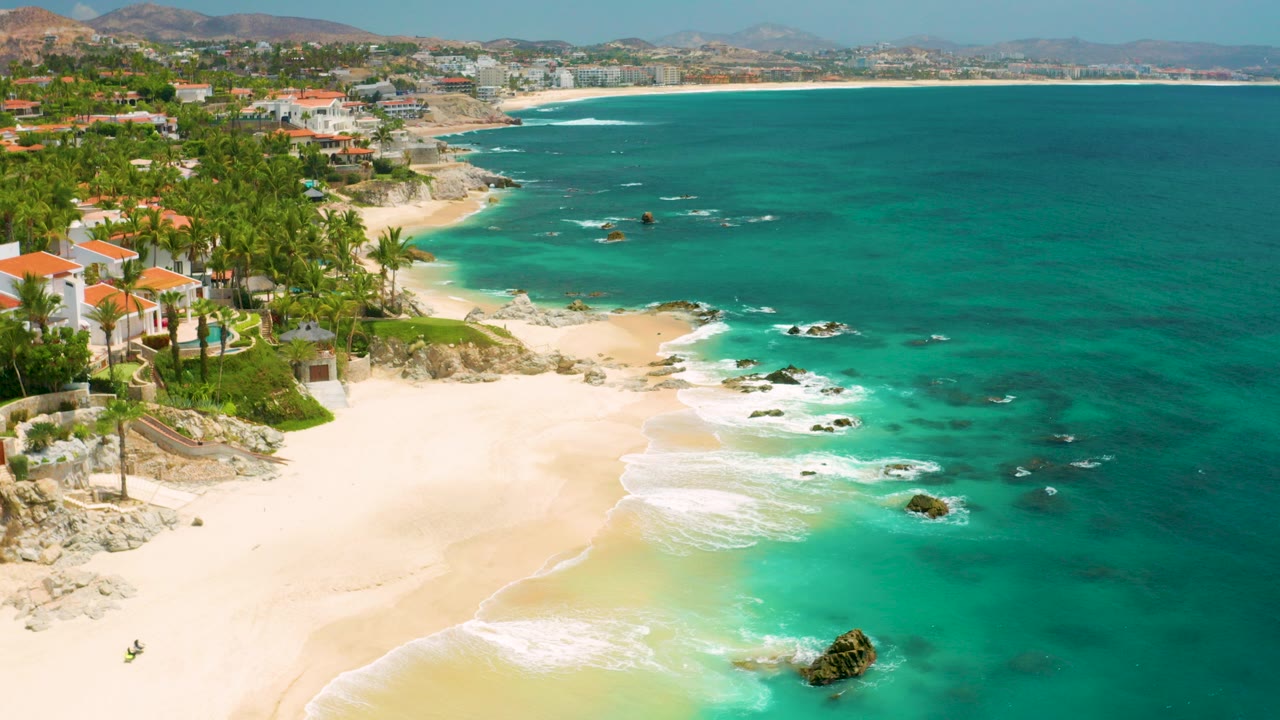 Aerial frame over Palmilla Beach Cabo, Mexico showing orange-roof beach houses and palms at left, pale sand below, turquoise surf around rocks, distant coast, hazy hills, and no visible people.