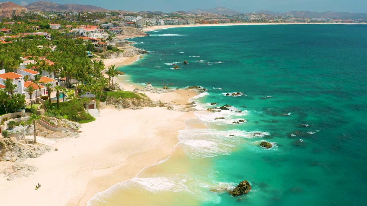 Wide aerial coastline with beach homes, palms, sand, rocks, and clear water.