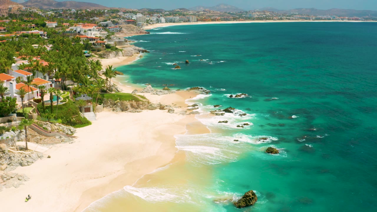 High aerial frame from Palmilla Beach Cabo, Mexico showing the sandy cove centered lower left, villas and palms along the bluff, rocks breaking the surf, broad teal water, and no visible people.