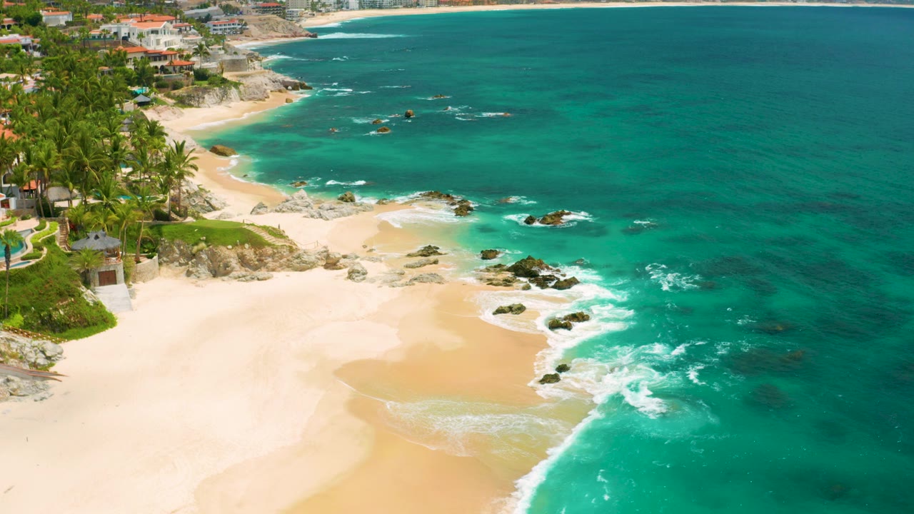 Opening aerial frame over Palmilla Beach Cabo, Mexico, with pale sand at lower left, turquoise surf around rocks, palms and coastal buildings along the upper shore, and no visible people.