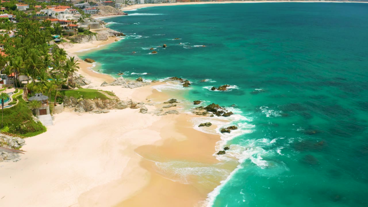 Second frame at Palmilla Beach Cabo shows a broad empty sand area, white foam crossing the shore, offshore rocks in blue-green water, and no visible people.