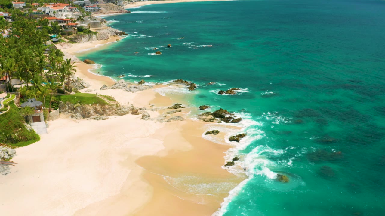 At 0:02, Palmilla Beach Cabo appears from above with more open water on the right, bright sand below, rocks in the surf line, beachfront buildings at the top, and no visible people.