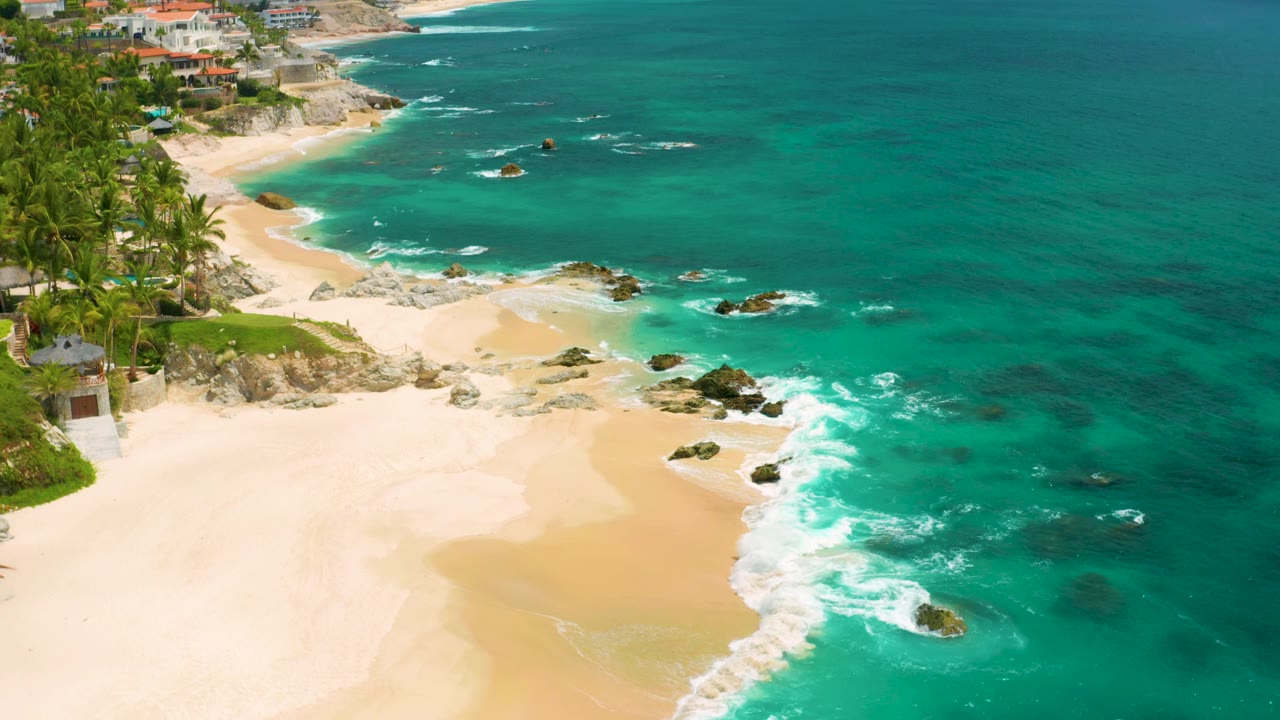 Aerial coastline with empty beach, rocky surf, palms, and clear ocean water.