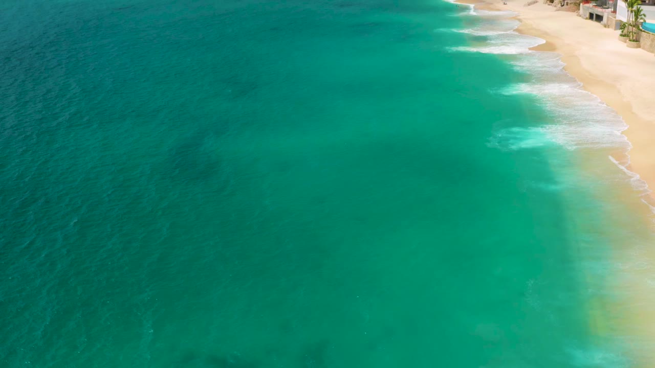 High aerial view over Palmilla Beach Cabo, Mexico, with turquoise water, darker reef patches below the surface, sand along the right edge, small surf, and no visible people.